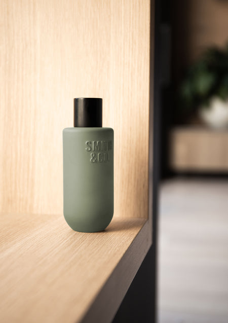 Sage green room spray at the edge of an oak shelf with a dark hallway blurred behind