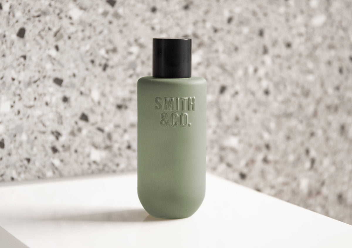 Landscape shot of the sage green room spray on a white bench against grey terrazzo
