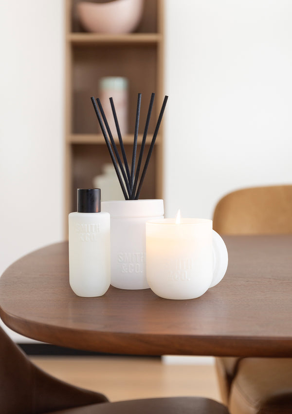 Room spray on a round timber table with pillar candles and a reed diffuser, shelving behind