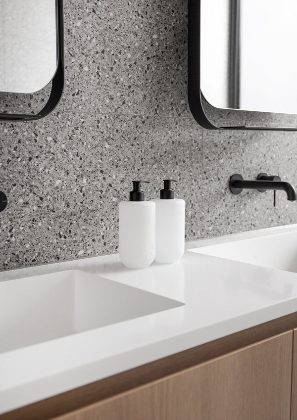 Smith & Co. pump bottles beside a white basin, with a terrazzo wall, round mirror and timber cabinet