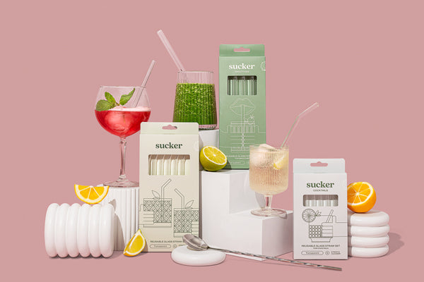 A collection of sustainable and reusable Sucker Cocktail Glass Drinking Straws - Transparent / Multi-coloured displayed alongside various drinks on a vibrant pink background.