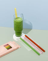 A pack of Reusable Glass Smoothie Straws - Transparent / Multi-coloured by Sucker next to a glass of green juice.