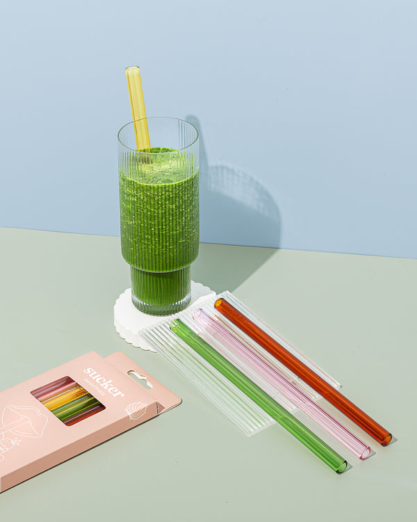 A pack of Reusable Glass Smoothie Straws - Transparent / Multi-coloured by Sucker next to a glass of green juice.