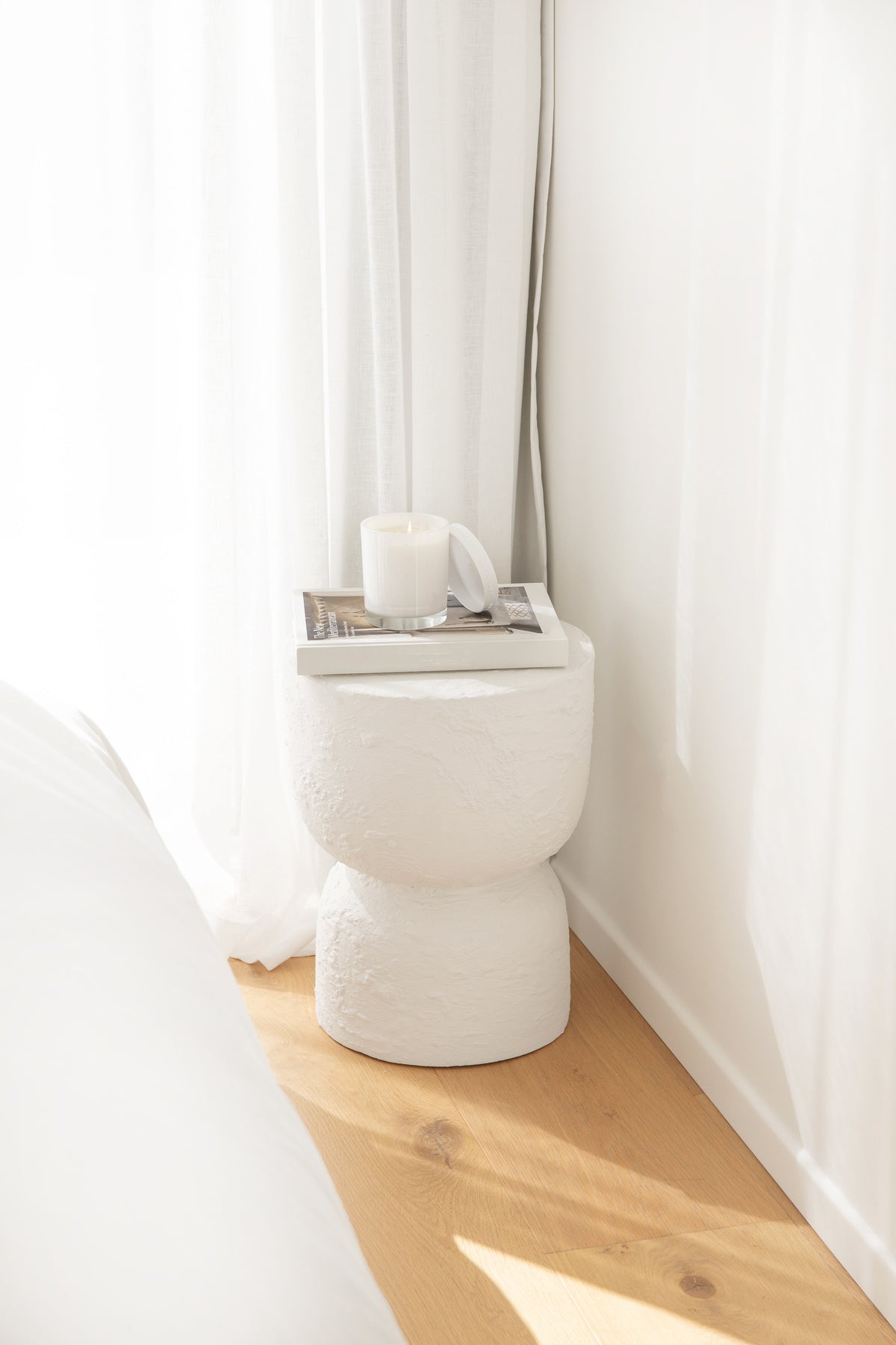 Naturals candle on a pedestal stool beside a bath, with a sheer curtain and timber floor