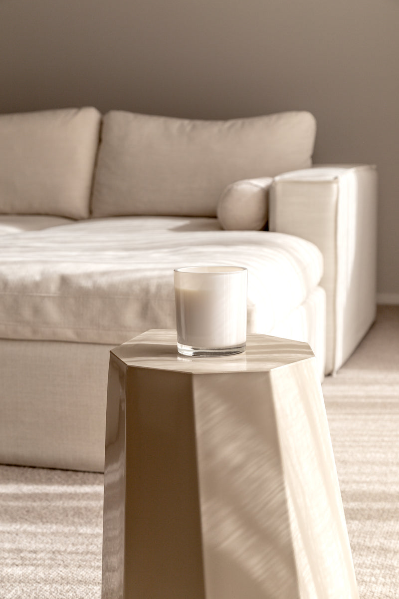 A minimalist creamy-toned living room with a warm soy candle styled on a sculptural beige side table, soft natural light casting calming shadows across a neutral sofa in the background.