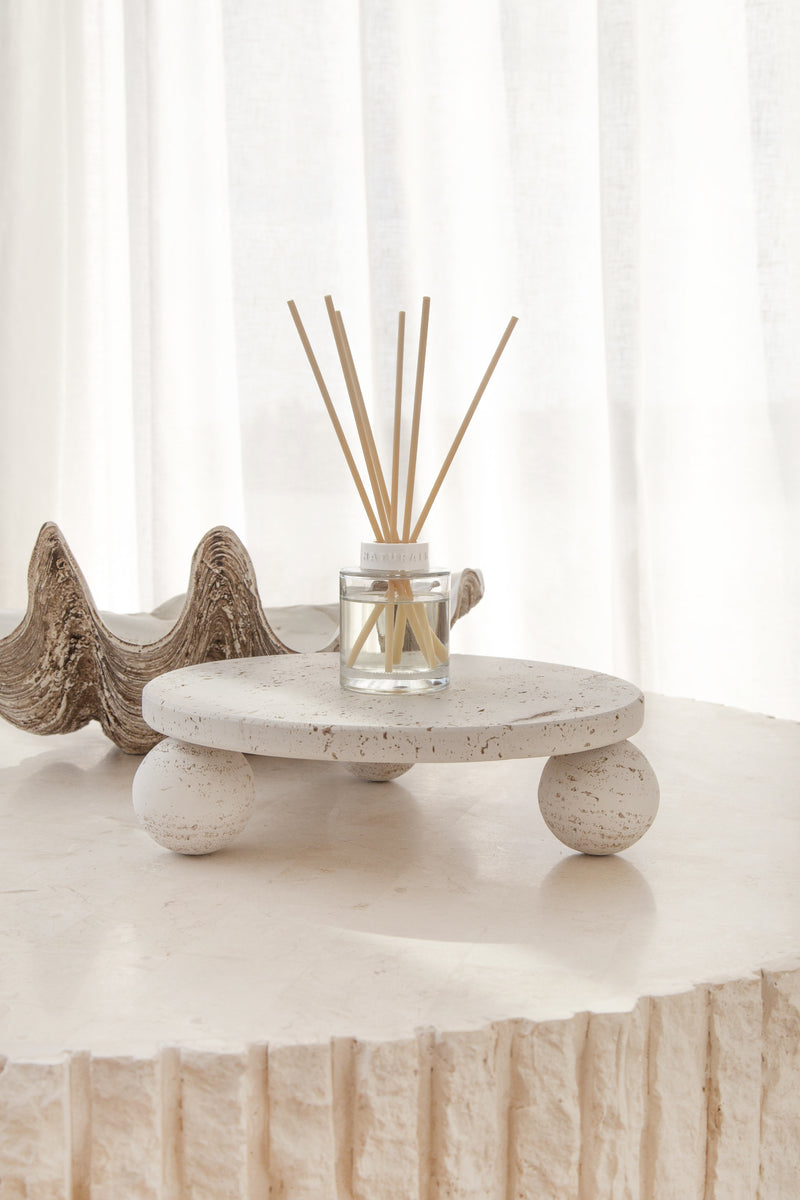 A minimalist Vanilla & Santal reed diffuser styled on a sculptural terrazzo platform, with natural rattan reeds fanning out beautifully against soft white curtains and a textured shell decor accent.