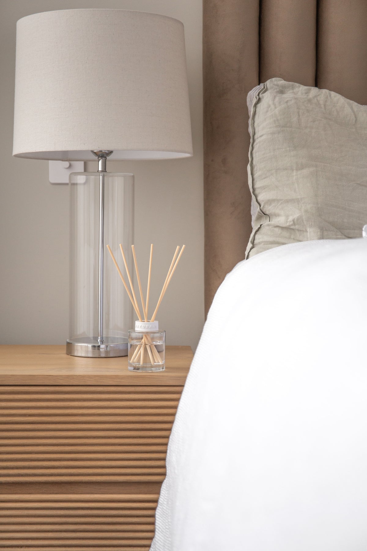Naturals diffuser on a fluted bedside table beside a tall glass lamp and white bedding
