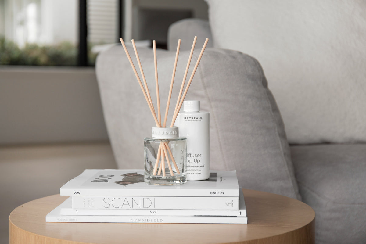 Naturals diffuser and a bottle on stacked books on a round table beside a grey sofa