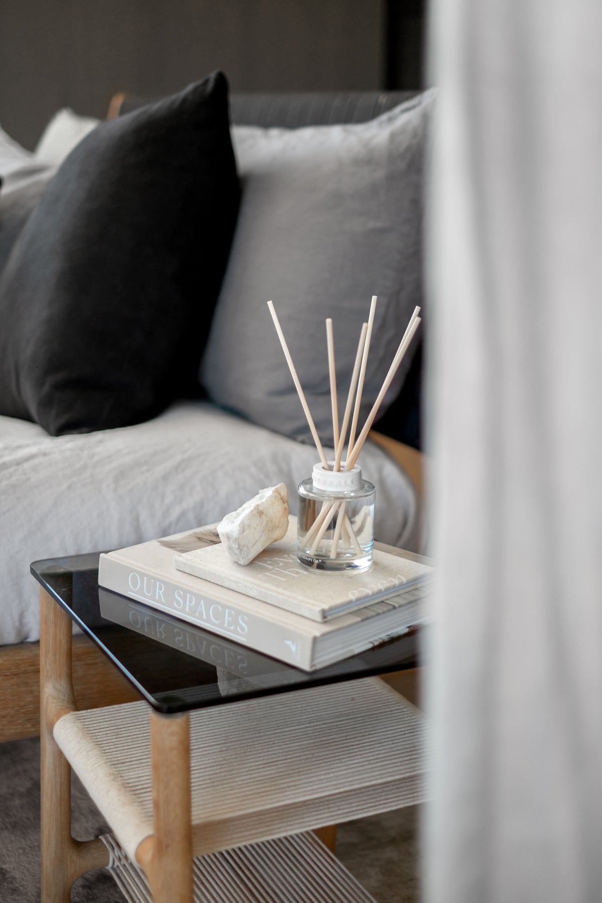Naturals diffuser and books on a glass-topped side table beside a bed with dark cushions