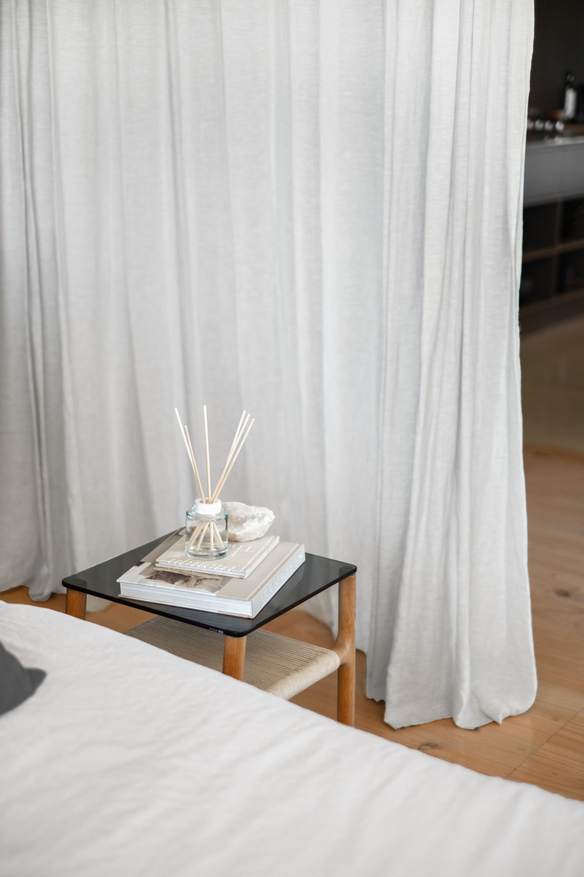 Naturals diffuser and books on a black side table beside a bed and sheer curtains