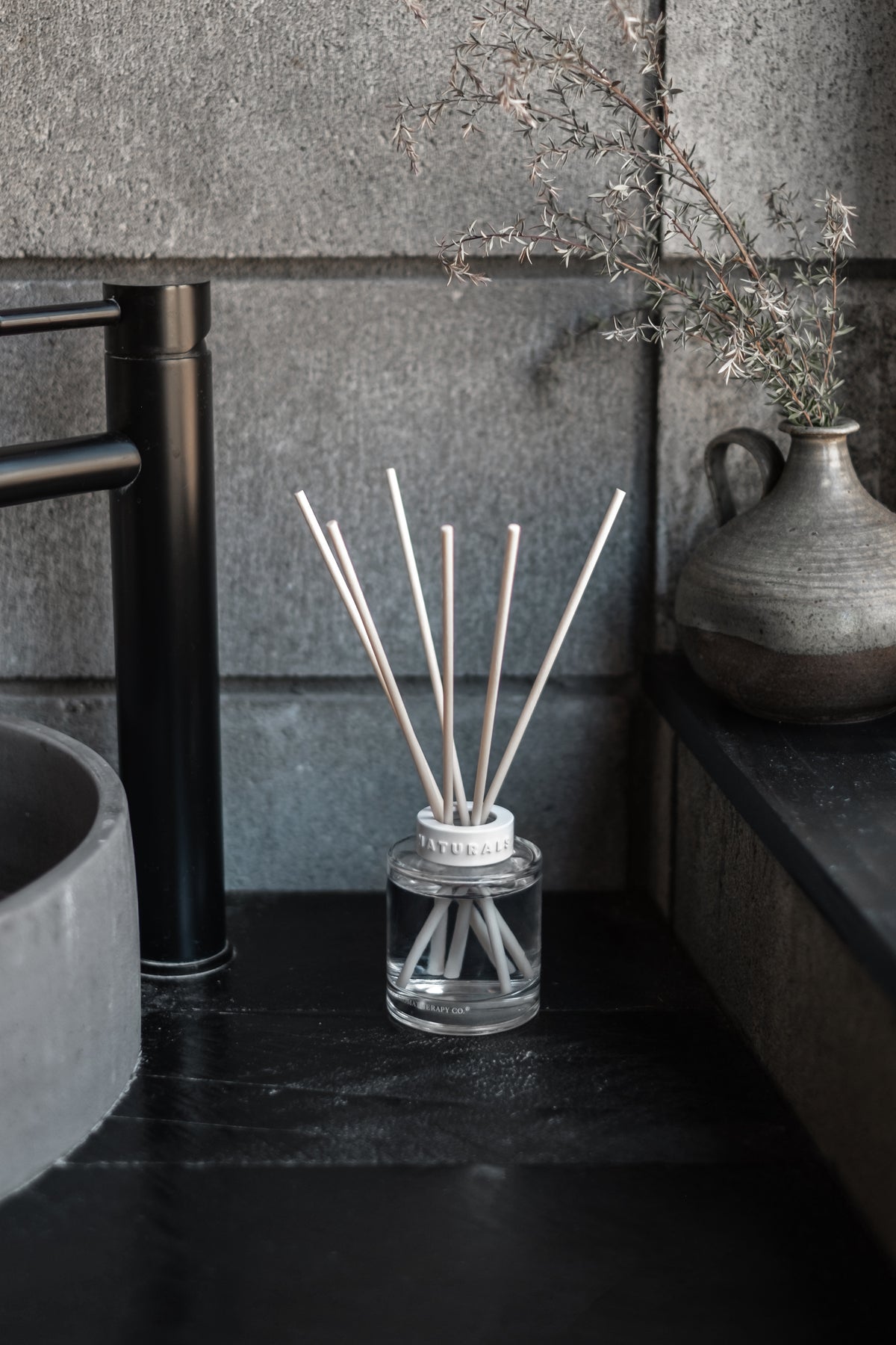 Naturals diffuser on a dark stone vanity with a black tap and a vase of dried flowers