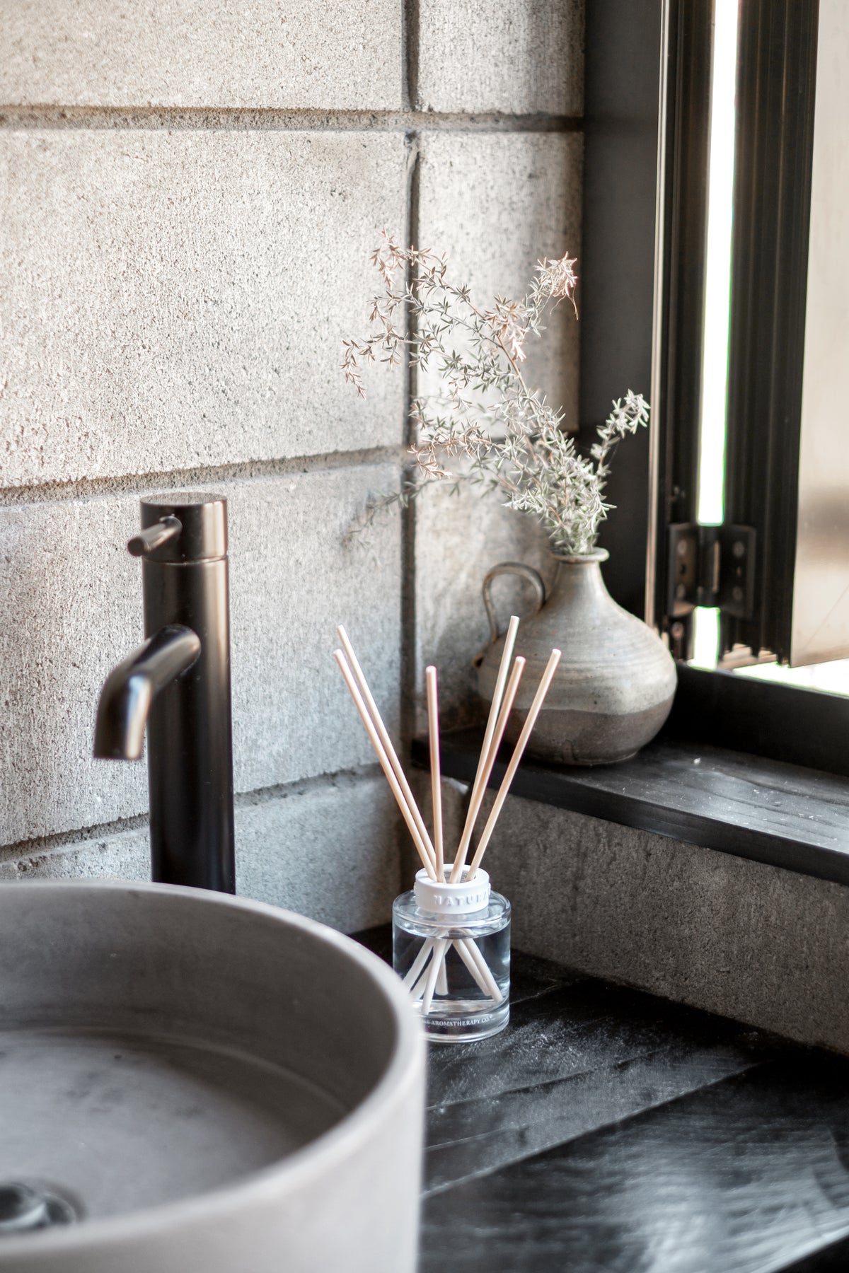 Naturals diffuser on a dark stone vanity with a black tap, basin and dried flowers