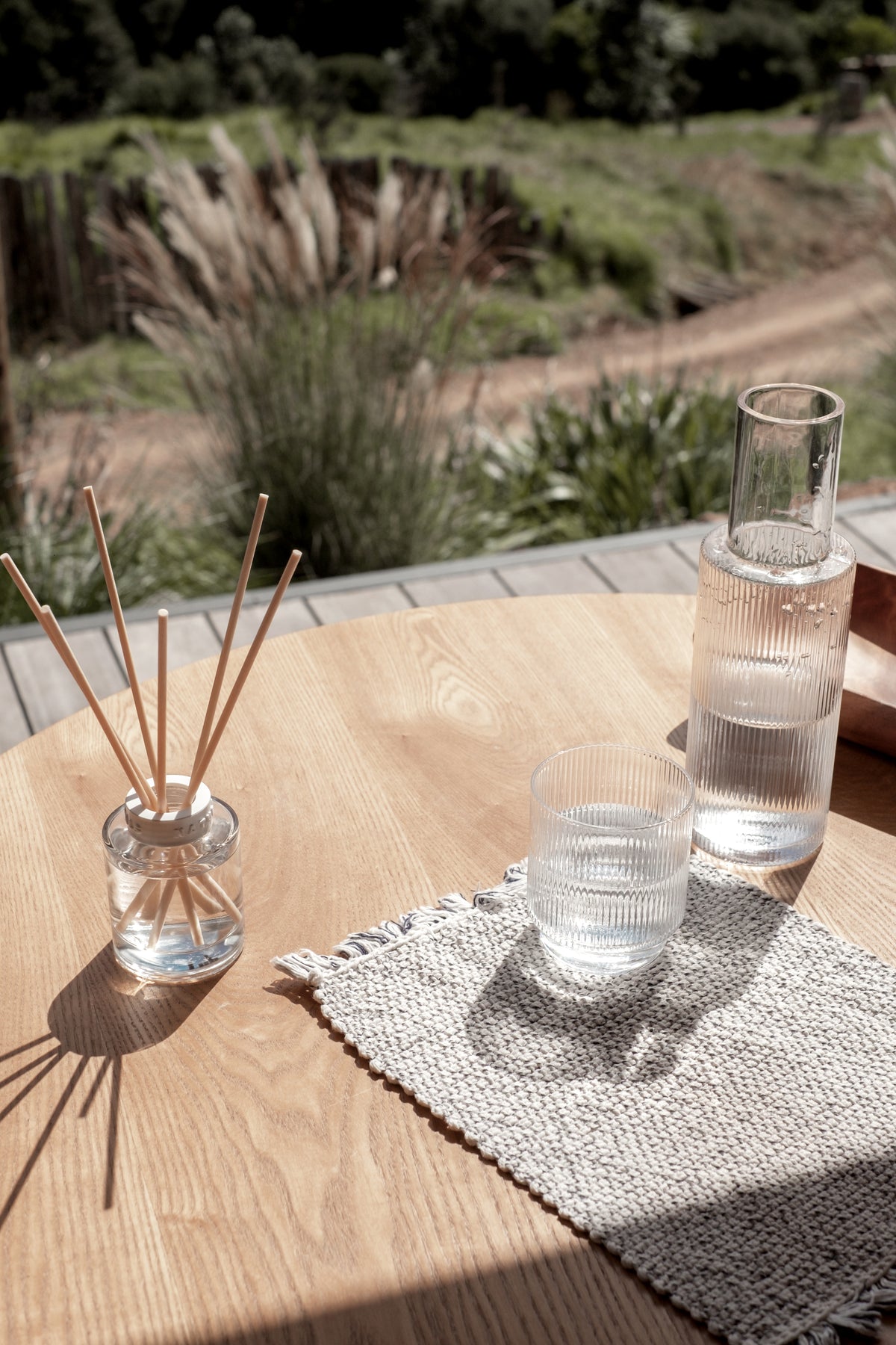 Naturals diffuser on an outdoor timber table with a carafe and glass, grasses beyond