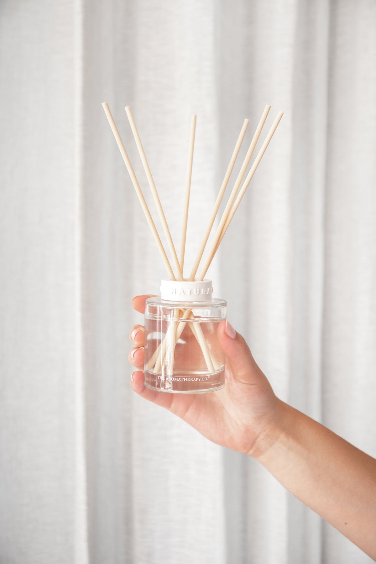 Hand holding the Naturals reed diffuser in front of a white curtain