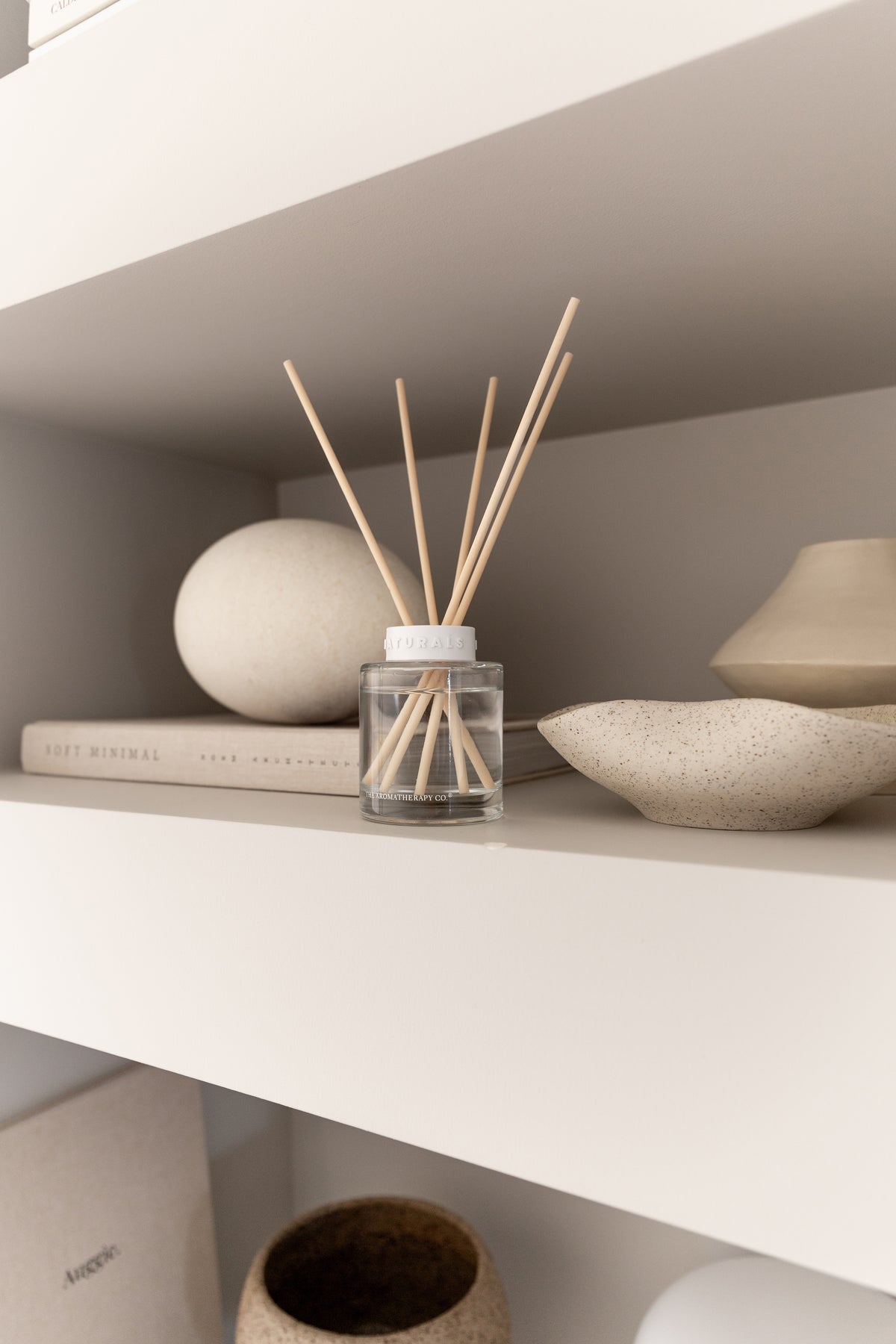 Naturals diffuser on a white shelf between a stone sphere and a shallow stone bowl