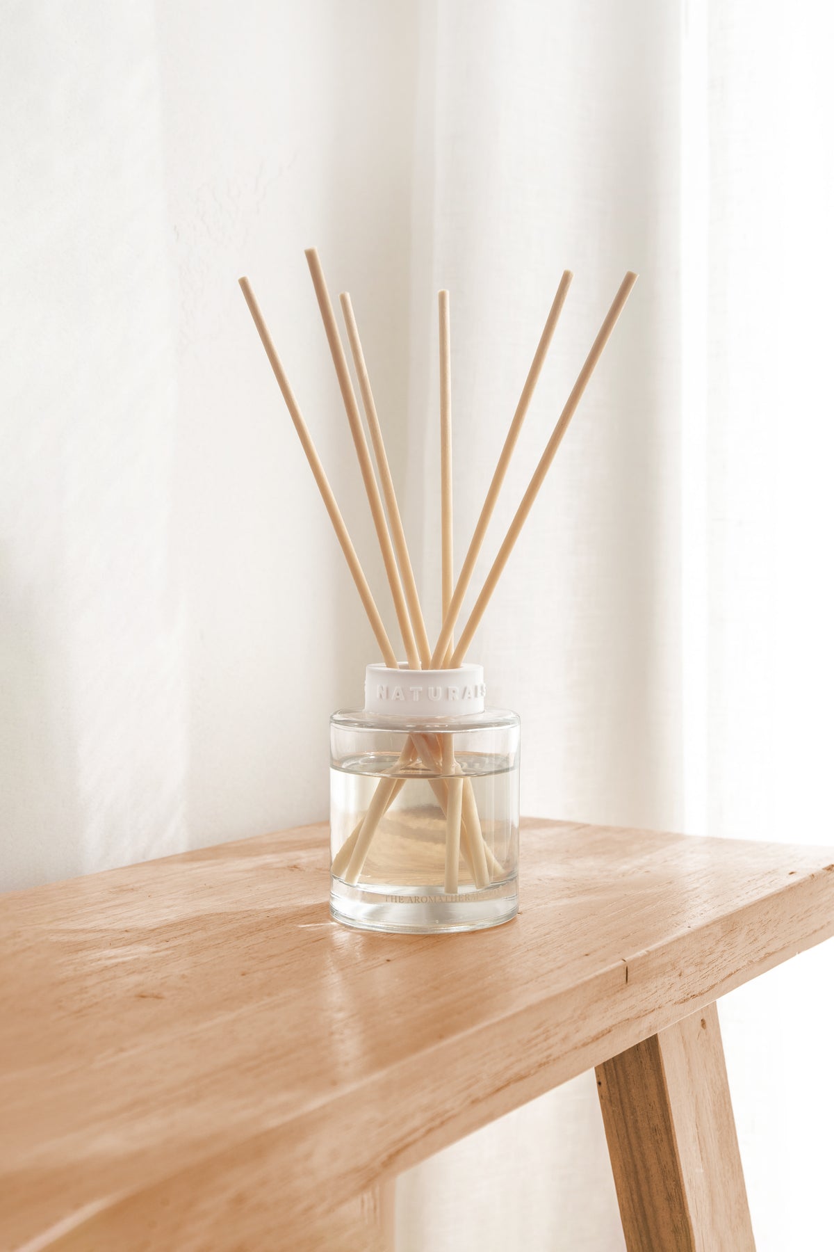 Naturals diffuser on the corner of a pale timber bench with a sheer curtain behind