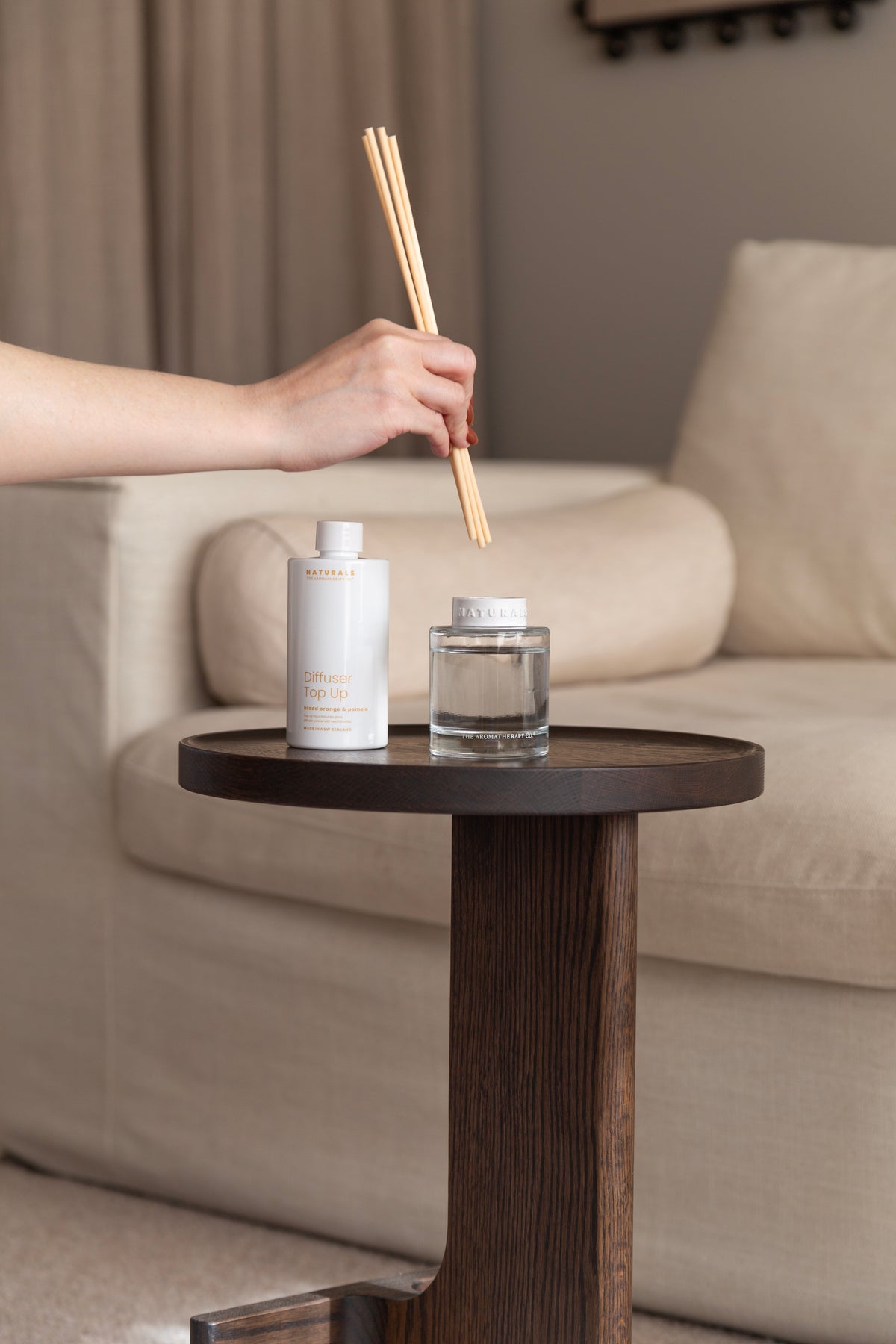 Hand holding the reeds above the Naturals diffuser and its refill bottle