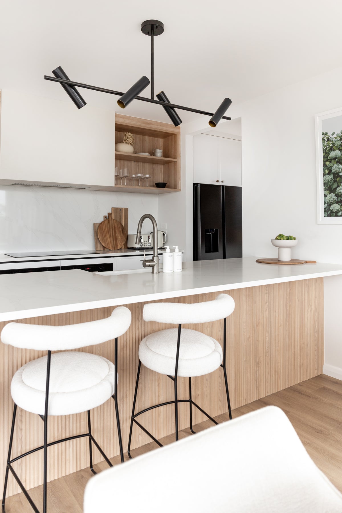 White kitchen with a black linear pendant, timber shelving and a fluted island, Naturals pump bottles on the benchtop