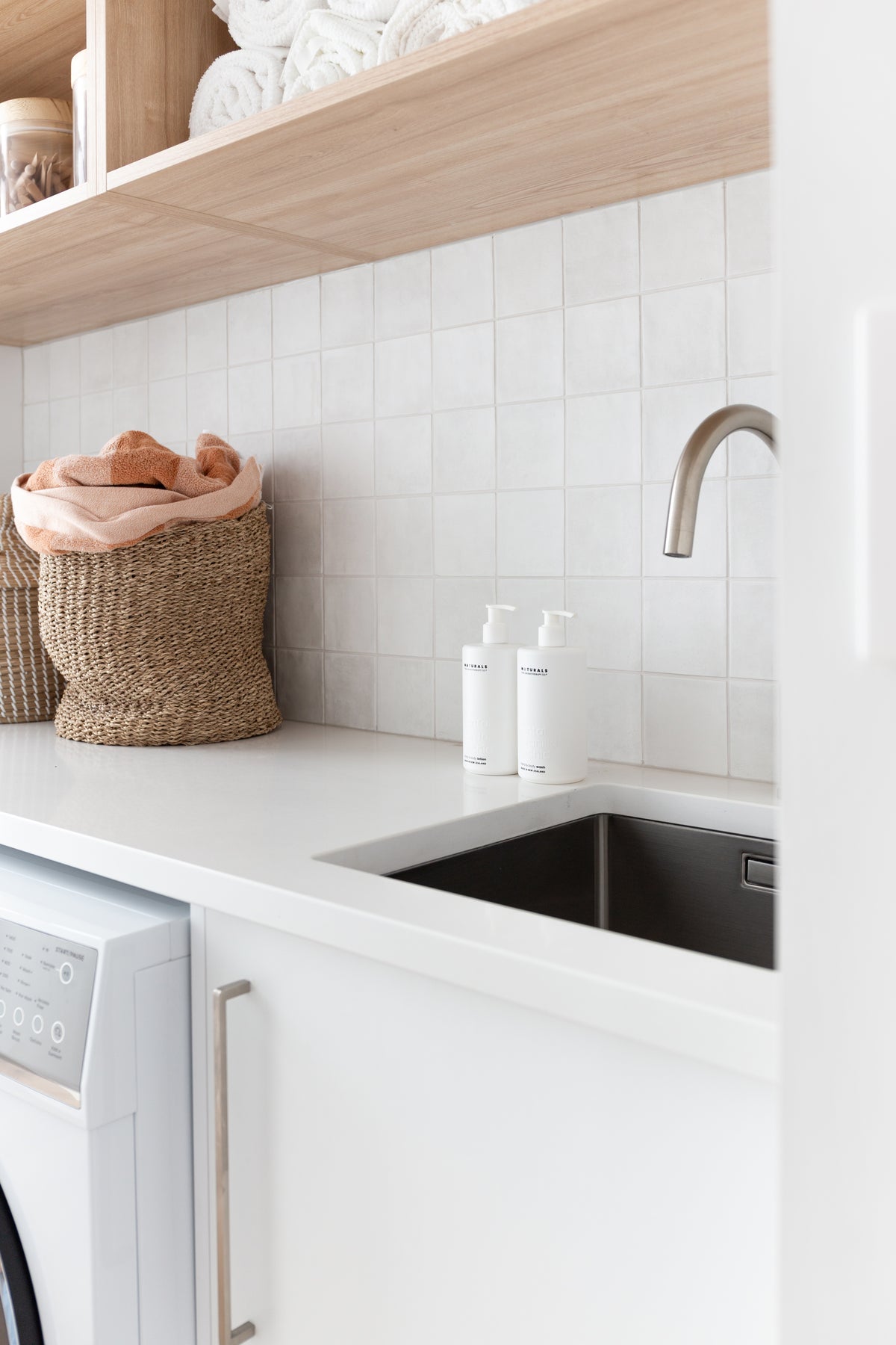 Laundry with folded white towels on a timber shelf, a woven basket and Naturals bottles by the sink