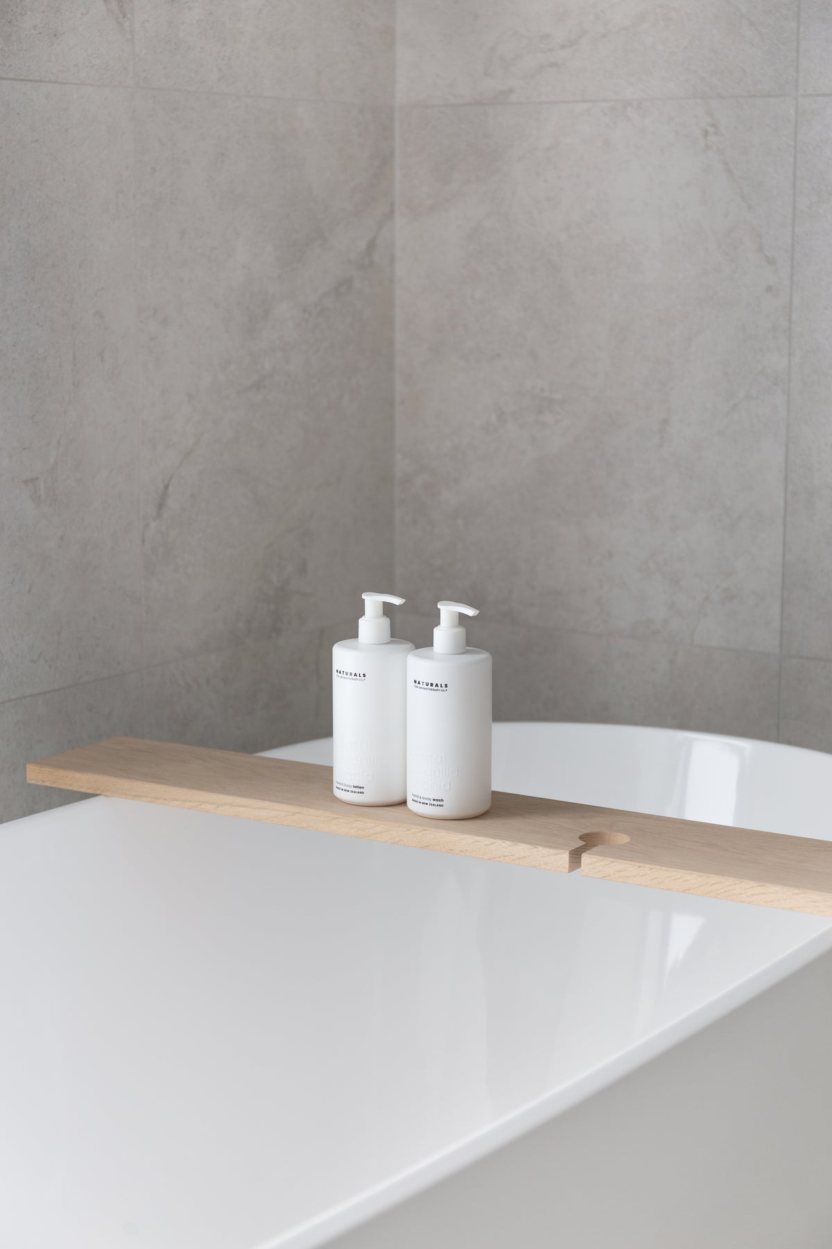 Freestanding bath with a timber bath board, two Naturals pump bottles resting on it against stone tiles