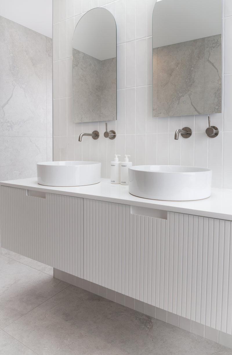 Modern minimalist bathroom with twin white vessel sinks, arched mirrors, and Vanilla & Santal Lotion and Wash bottles on a fluted vanity bench — styled in a soft neutral palette with brushed nickel fixtures.