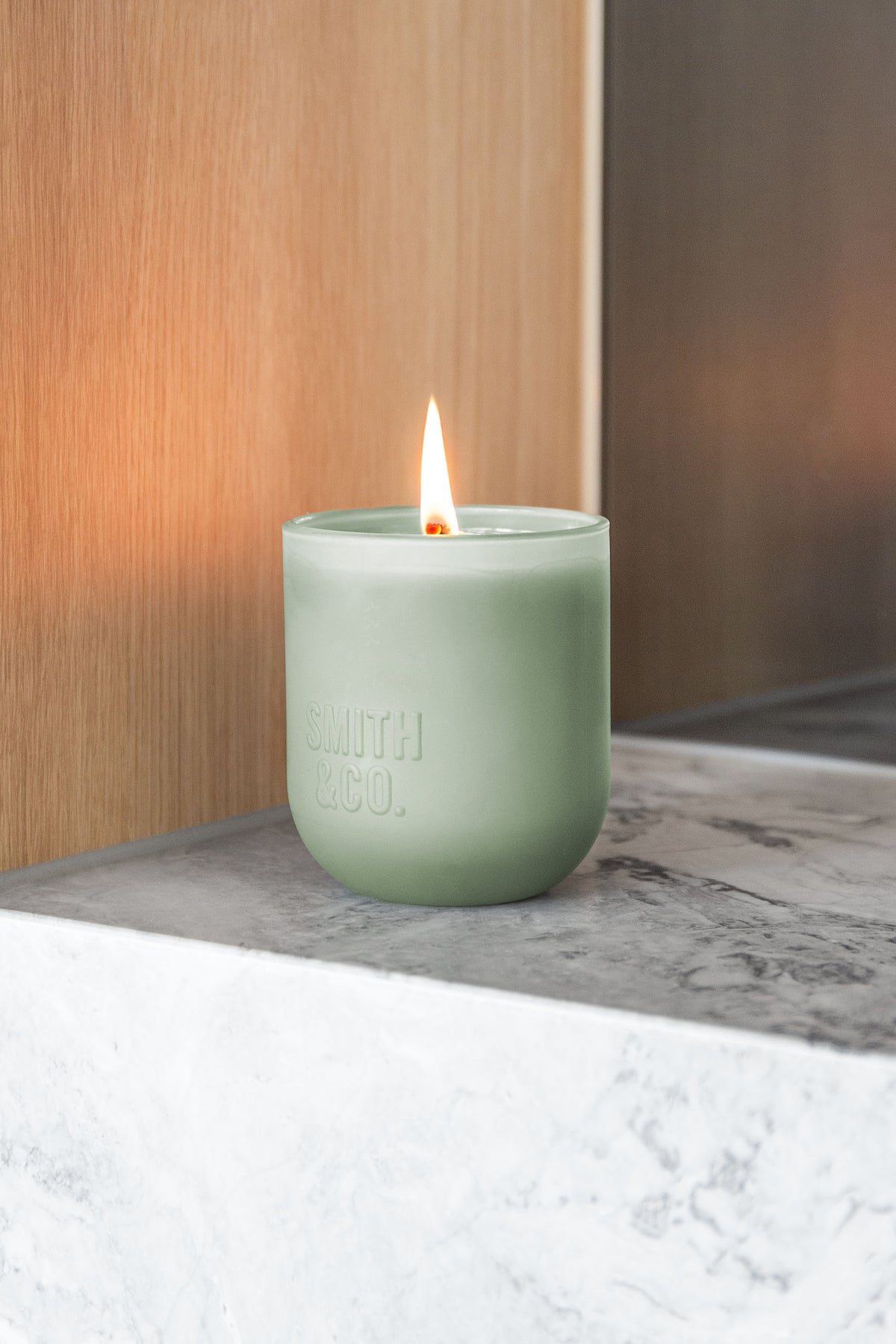 Lit Smith and Co candle on a marble ledge in front of a pale timber panel
