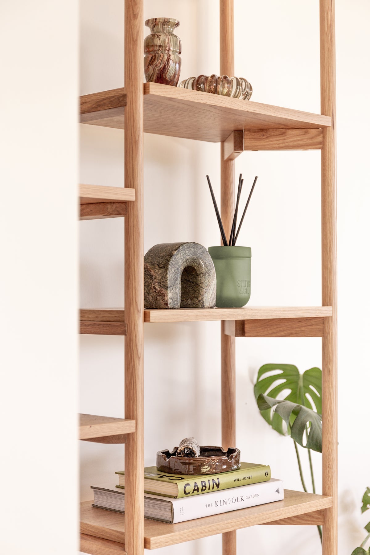 Smith and Co reed diffuser and a stone arch on an oak shelf above a monstera and books