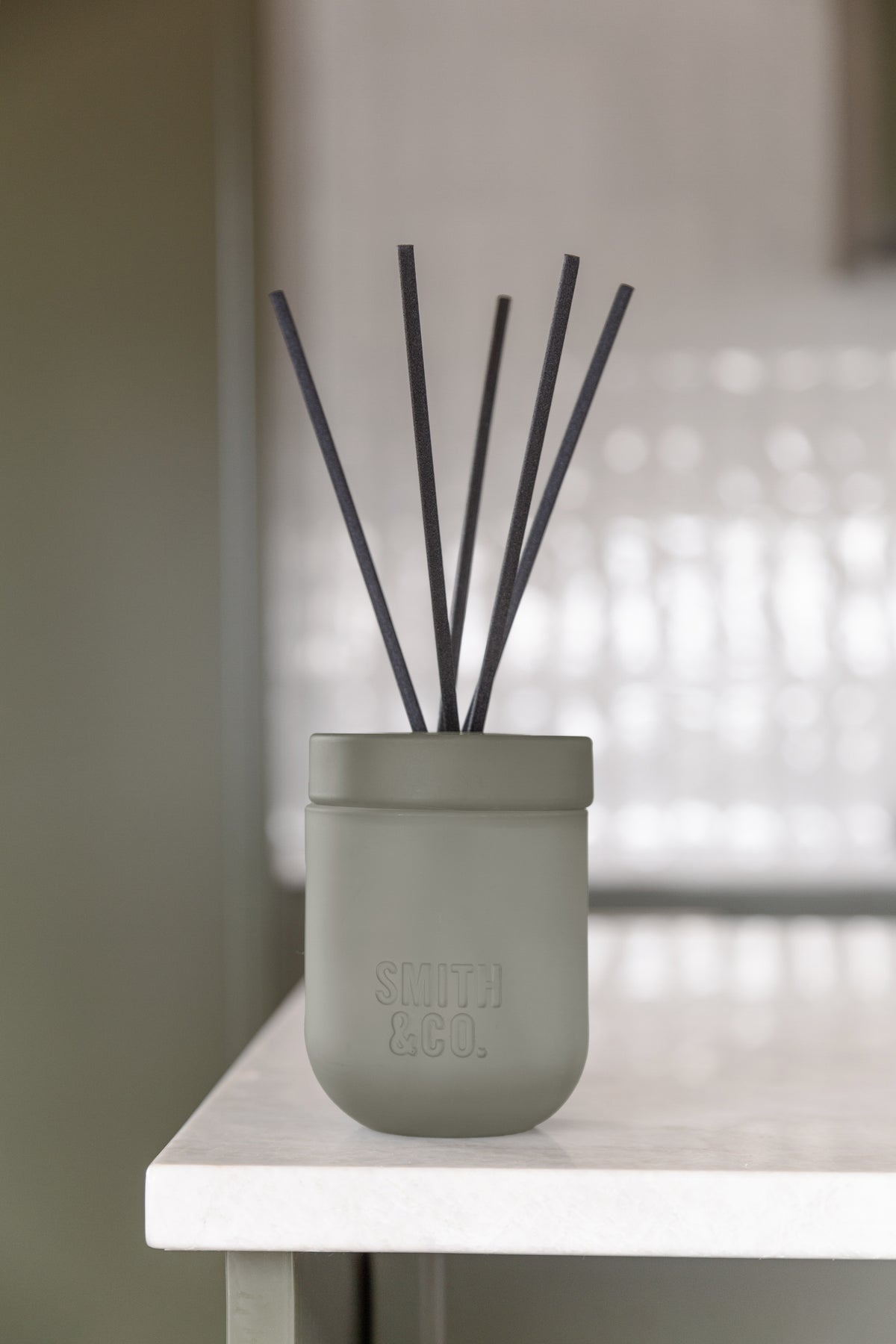Smith and Co reed diffuser on the corner of a white table in front of textured glass