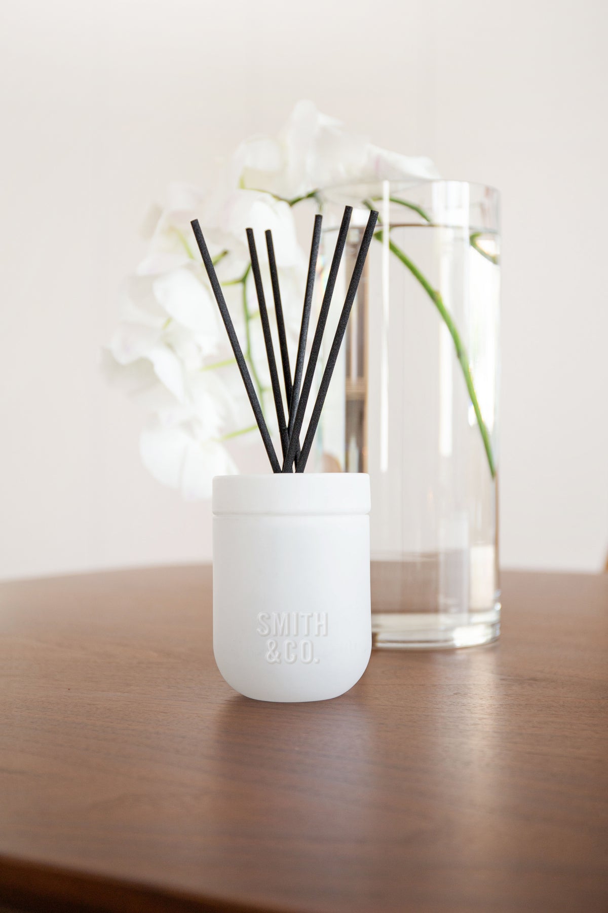 White Smith and Co diffuser beside a glass vase of white flowers on a dark table