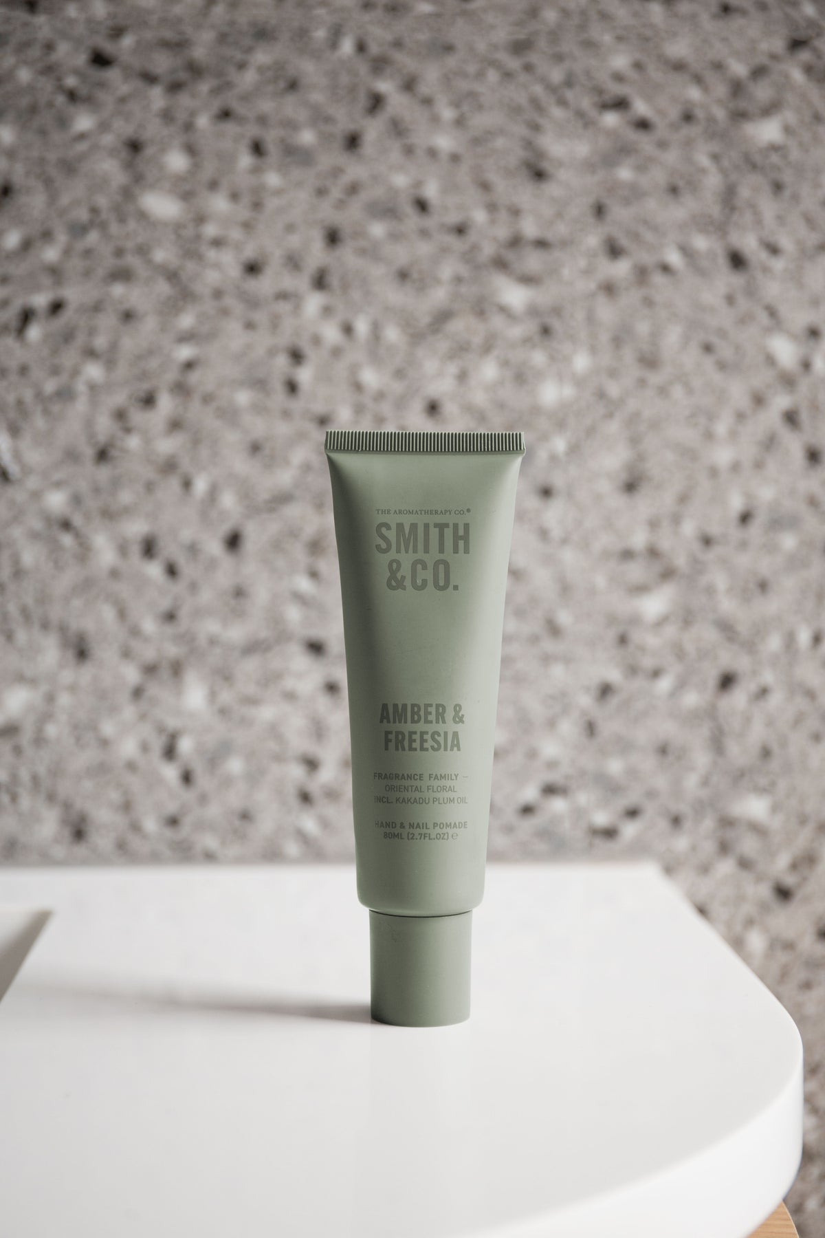 Hand cream tube standing on a curved white shelf against a speckled terrazzo wall