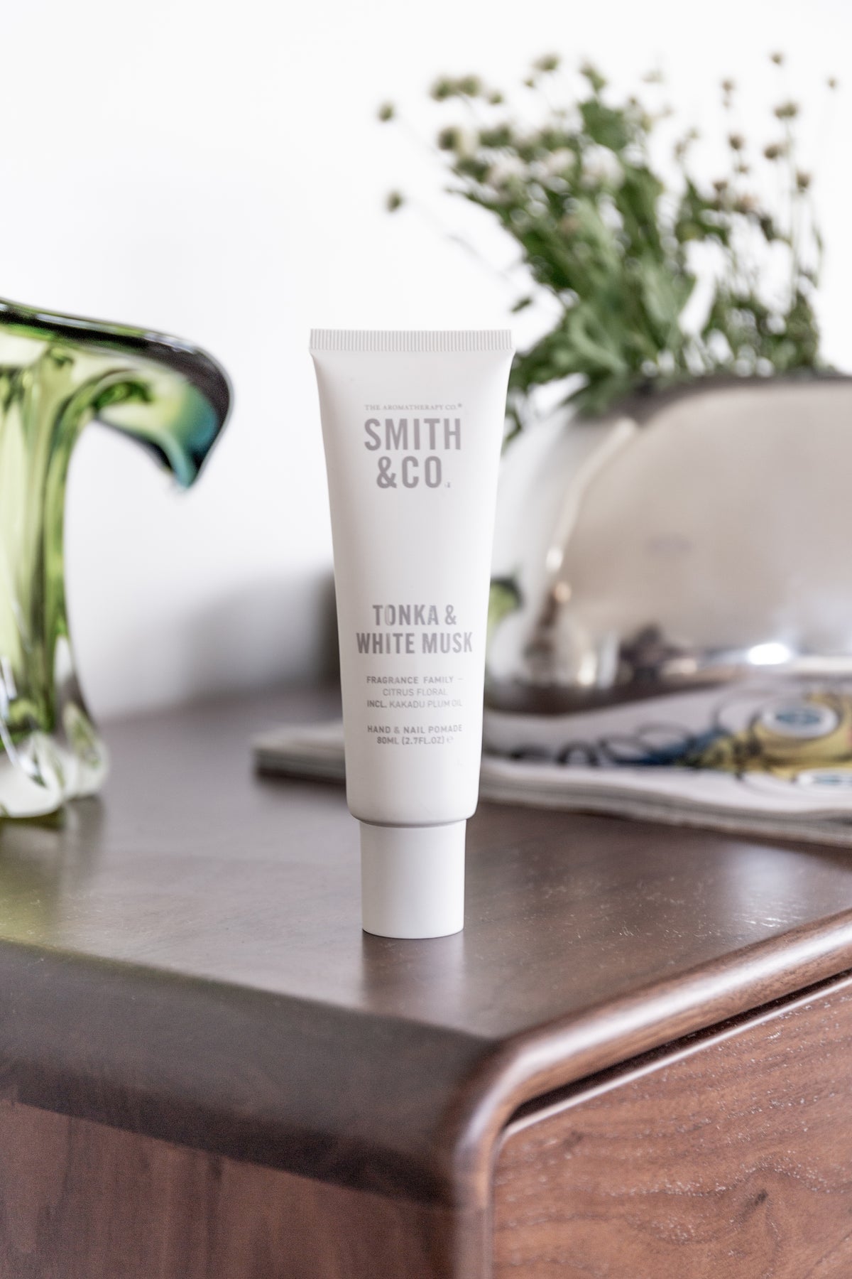 Hand cream on a timber vanity beside green glassware and a vase of dried flowers