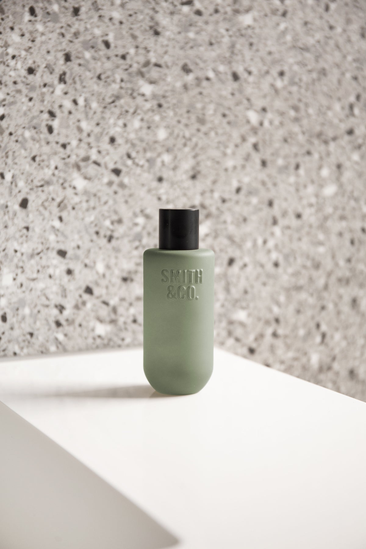 Room spray on a white bench corner with a speckled terrazzo wall behind, portrait view