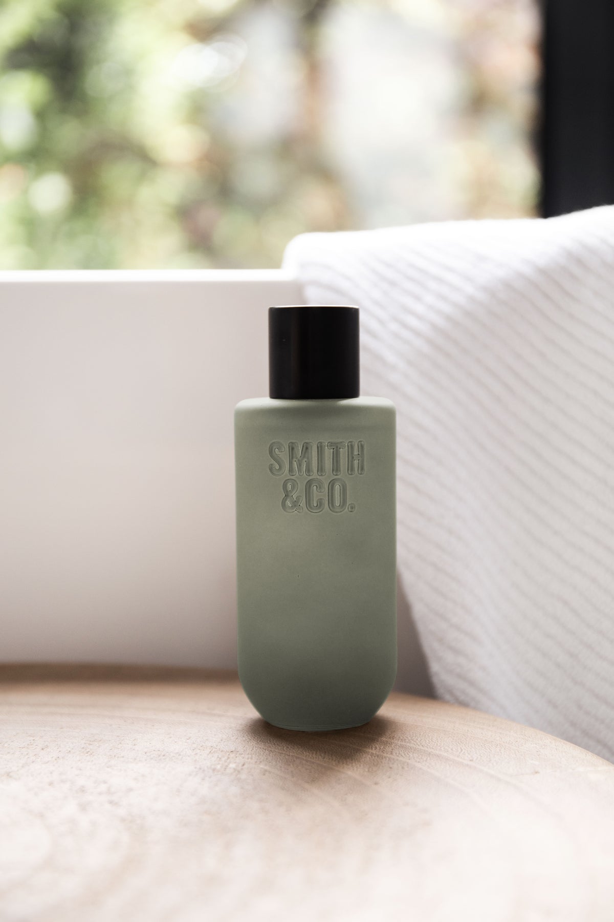 Sage green room spray on a round timber stool beside a folded white towel by a window