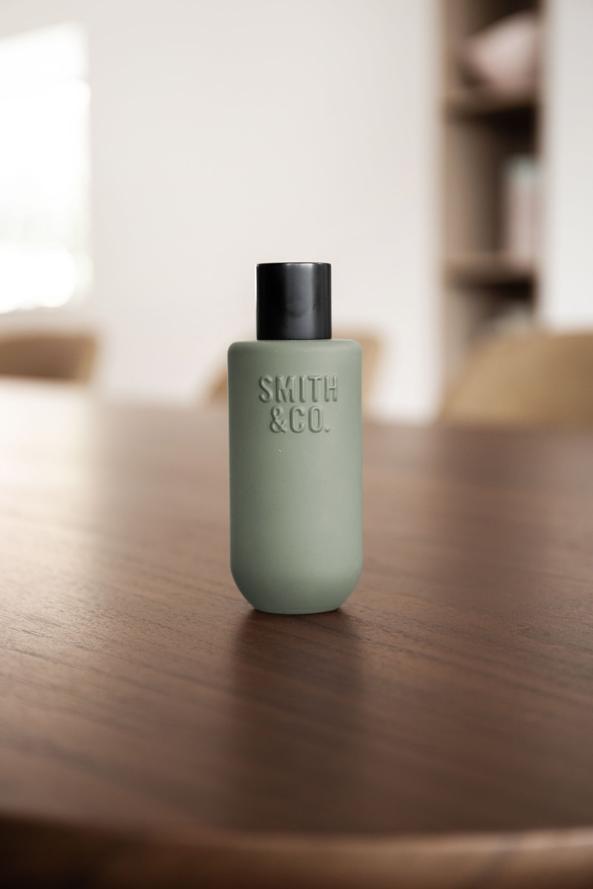 Front view of the sage green spray bottle on a walnut dining table with shelving blurred behind