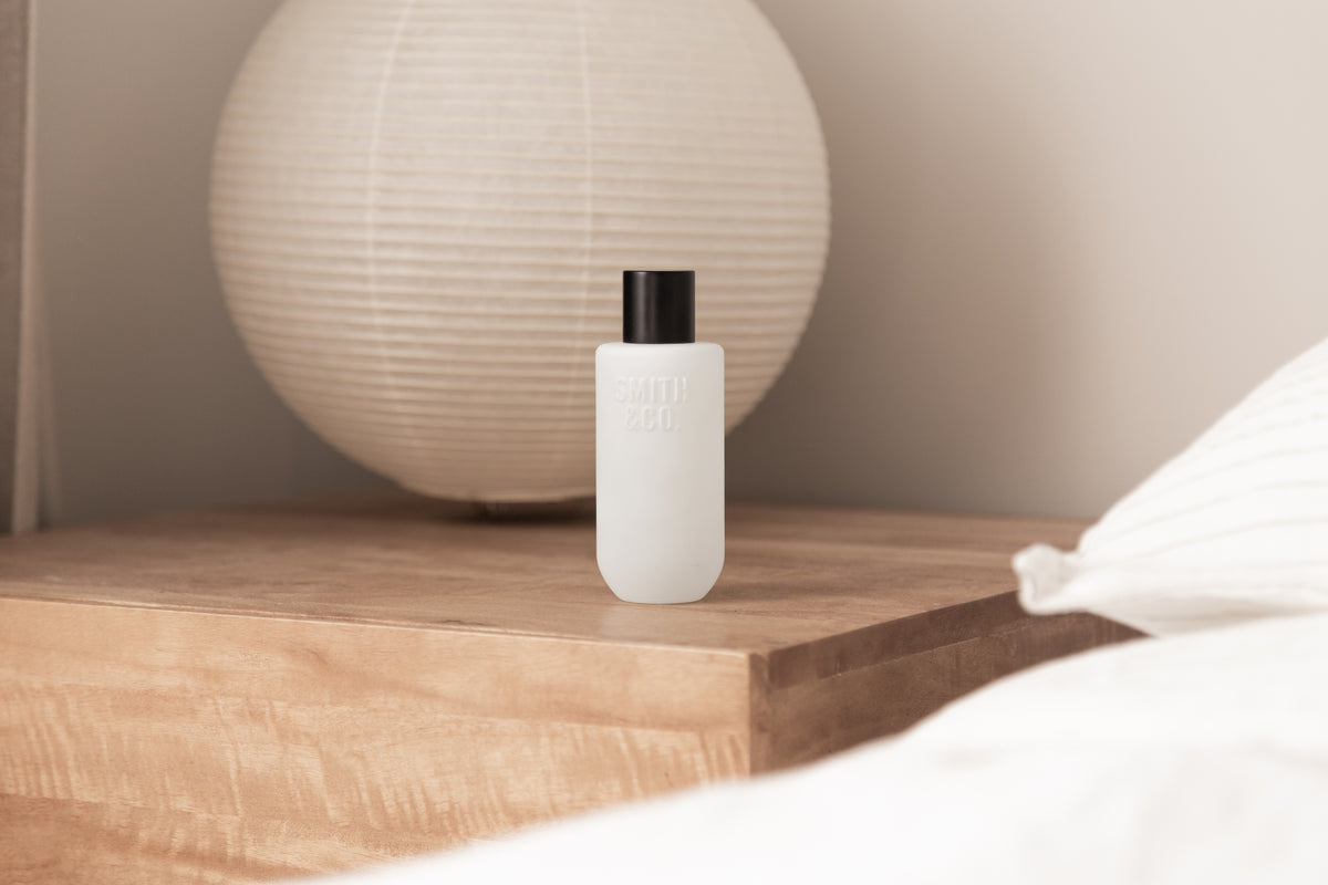Room spray on a timber bedside table next to a round paper lamp and white bedding