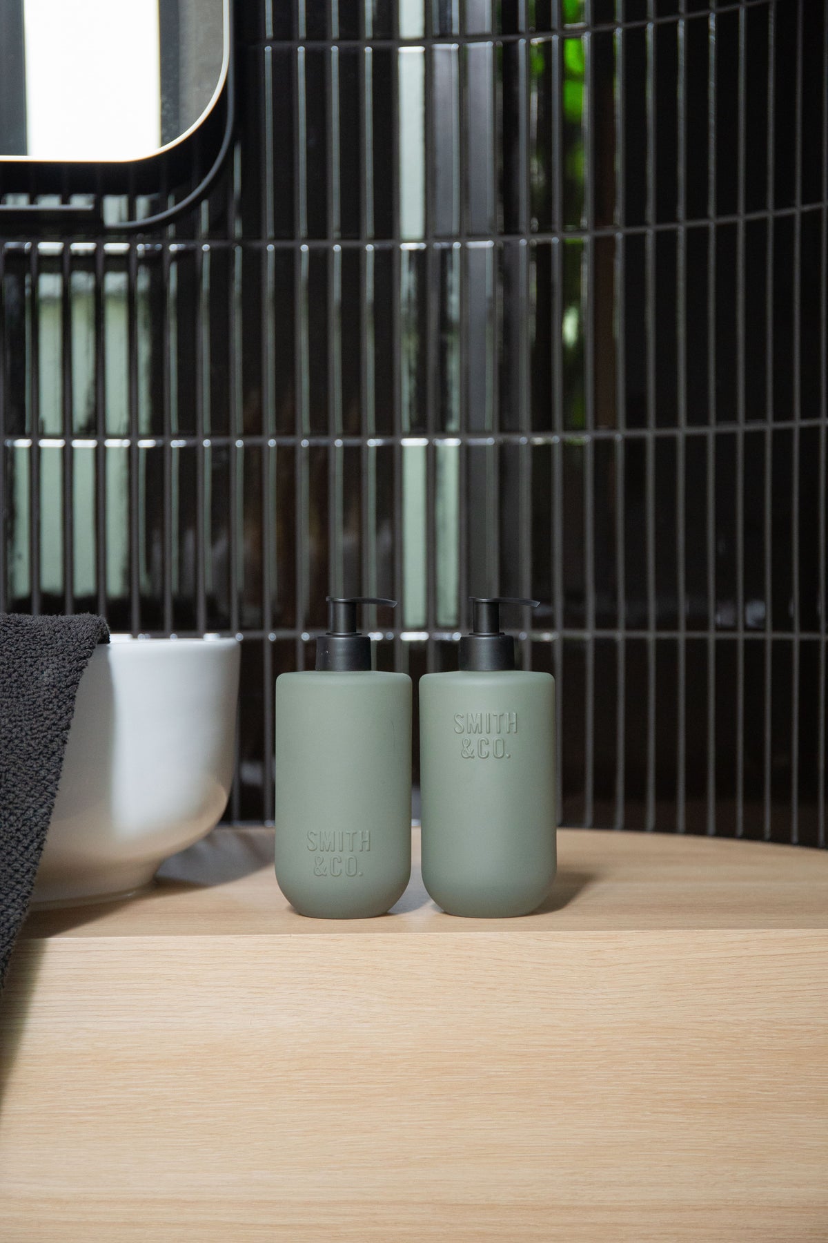 Two sage Smith & Co. bottles on a timber bench against dark green vertical tiles, close view