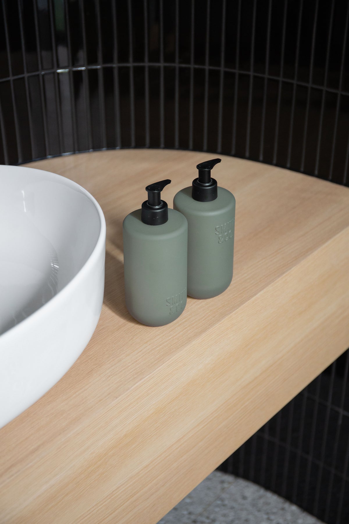 Two sage Smith & Co. bottles on a timber bench beside a white vessel basin with black tiles