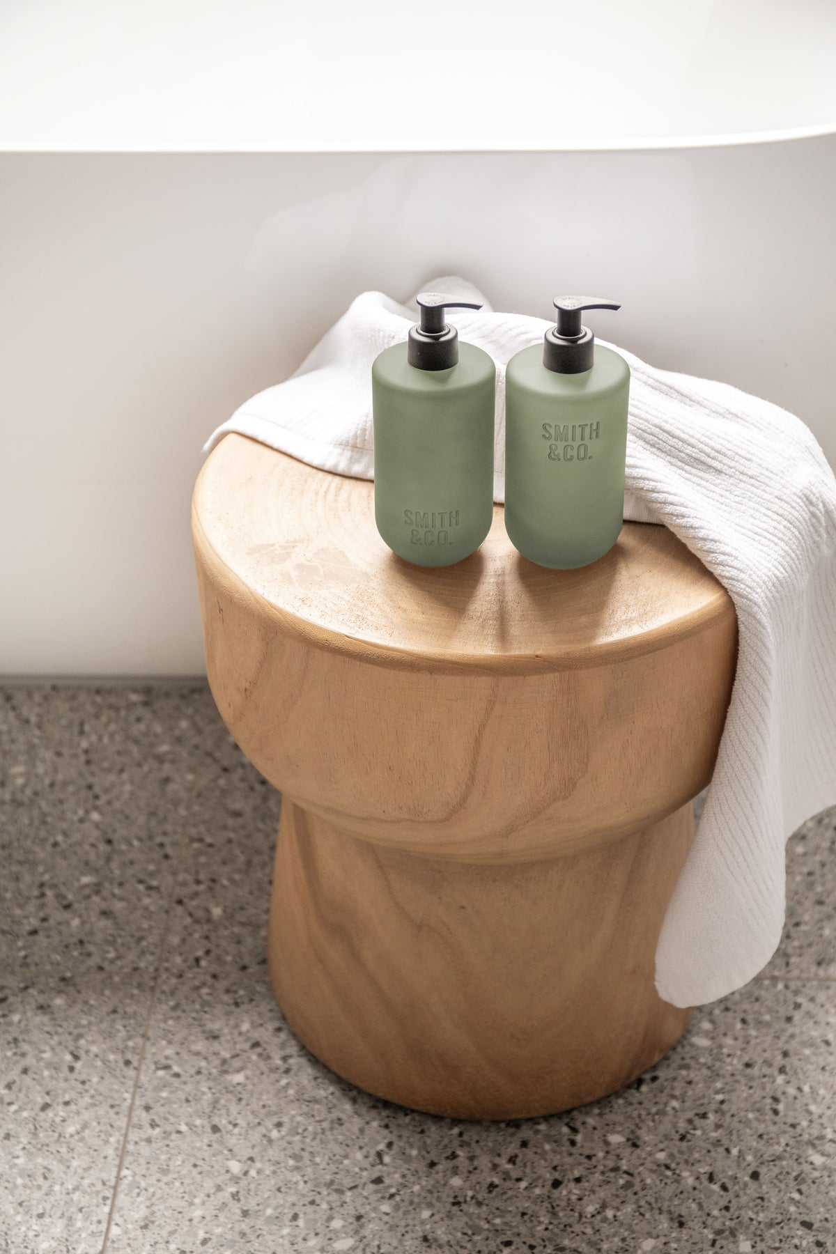 Two sage green bottles on a round timber stool with a white towel