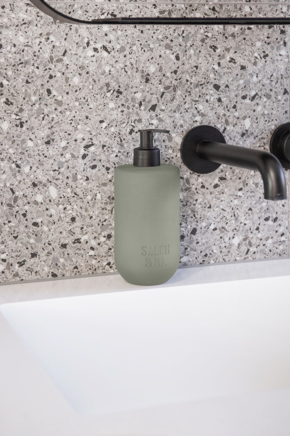 Sage green bottles on a terrazzo shelf beside black tapware