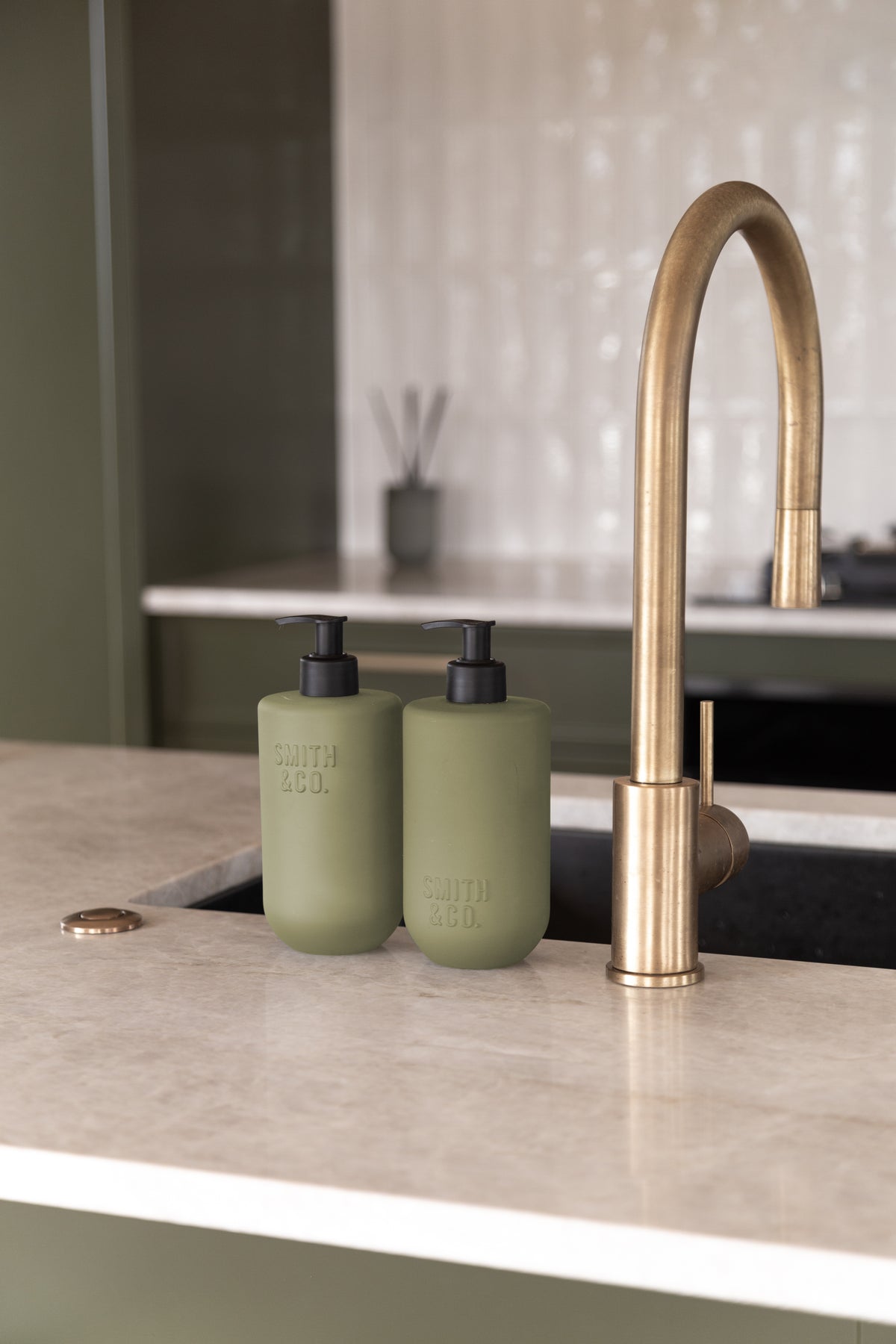 Sage green bottles by a brass tap against white tiles