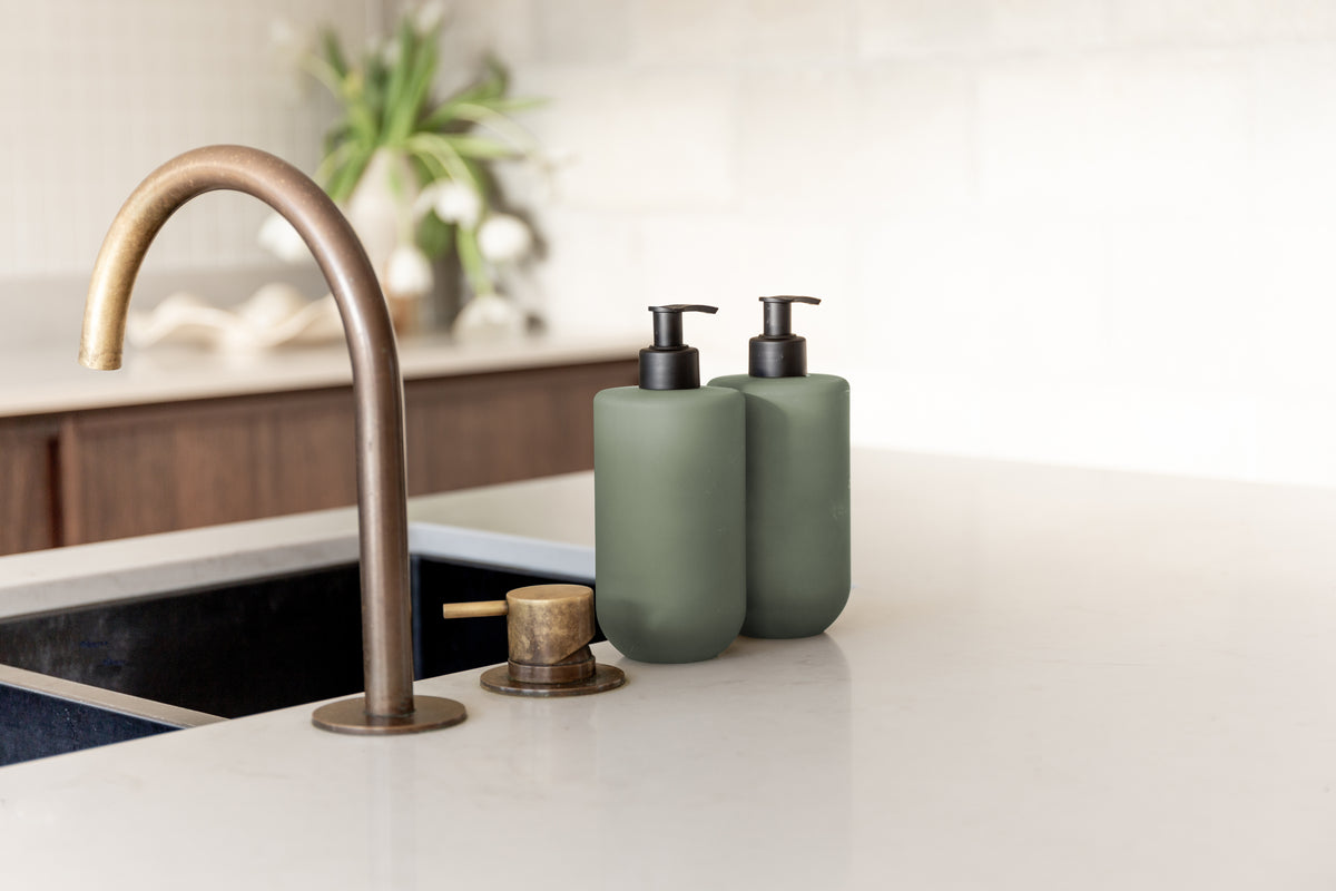 Two sage Smith & Co. bottles beside a brass mixer and black sink, with a plant behind