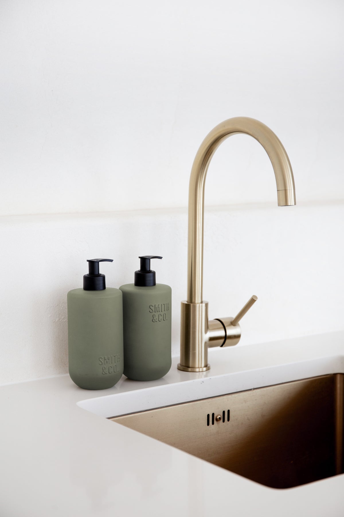 Two sage Smith & Co. bottles beside a brass gooseneck mixer and brass kitchen sink