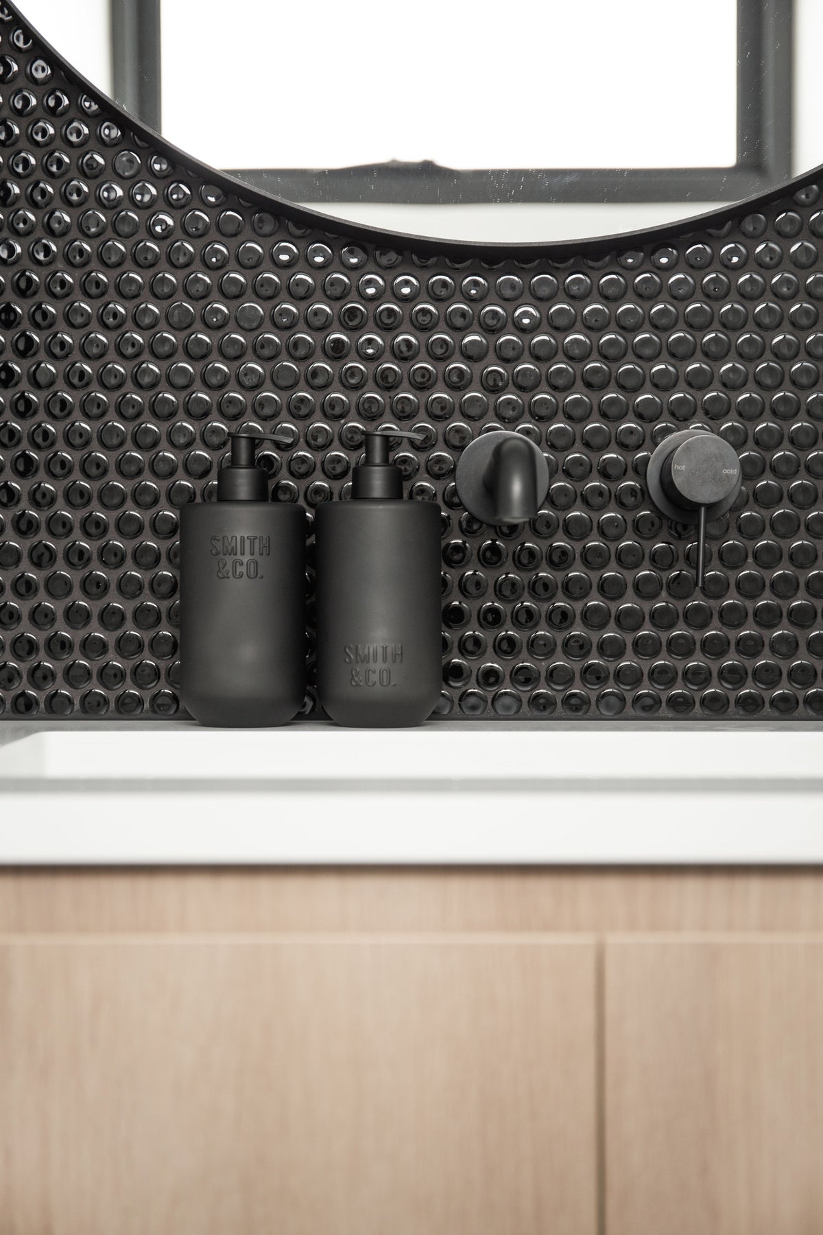 Smith & Co. bottles on a white basin with black penny round tiles and a timber vanity below