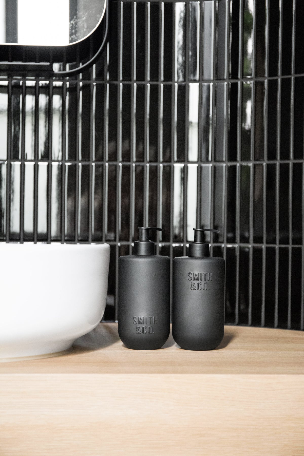 Two Smith & Co. bottles on a timber vanity beside a basin, against glossy black tiles