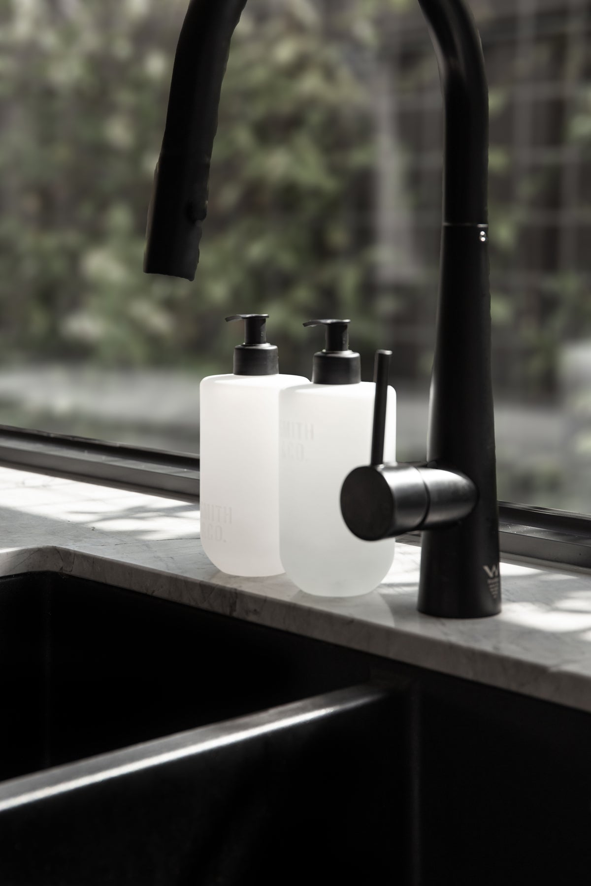 Frosted bottles beside a black tap at a kitchen sink with a city window behind