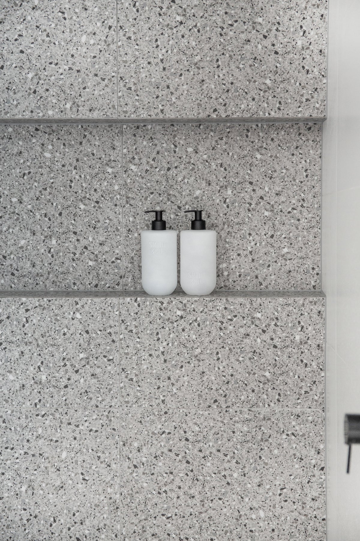 Two frosted pump bottles on a narrow ledge set into a grey terrazzo wall
