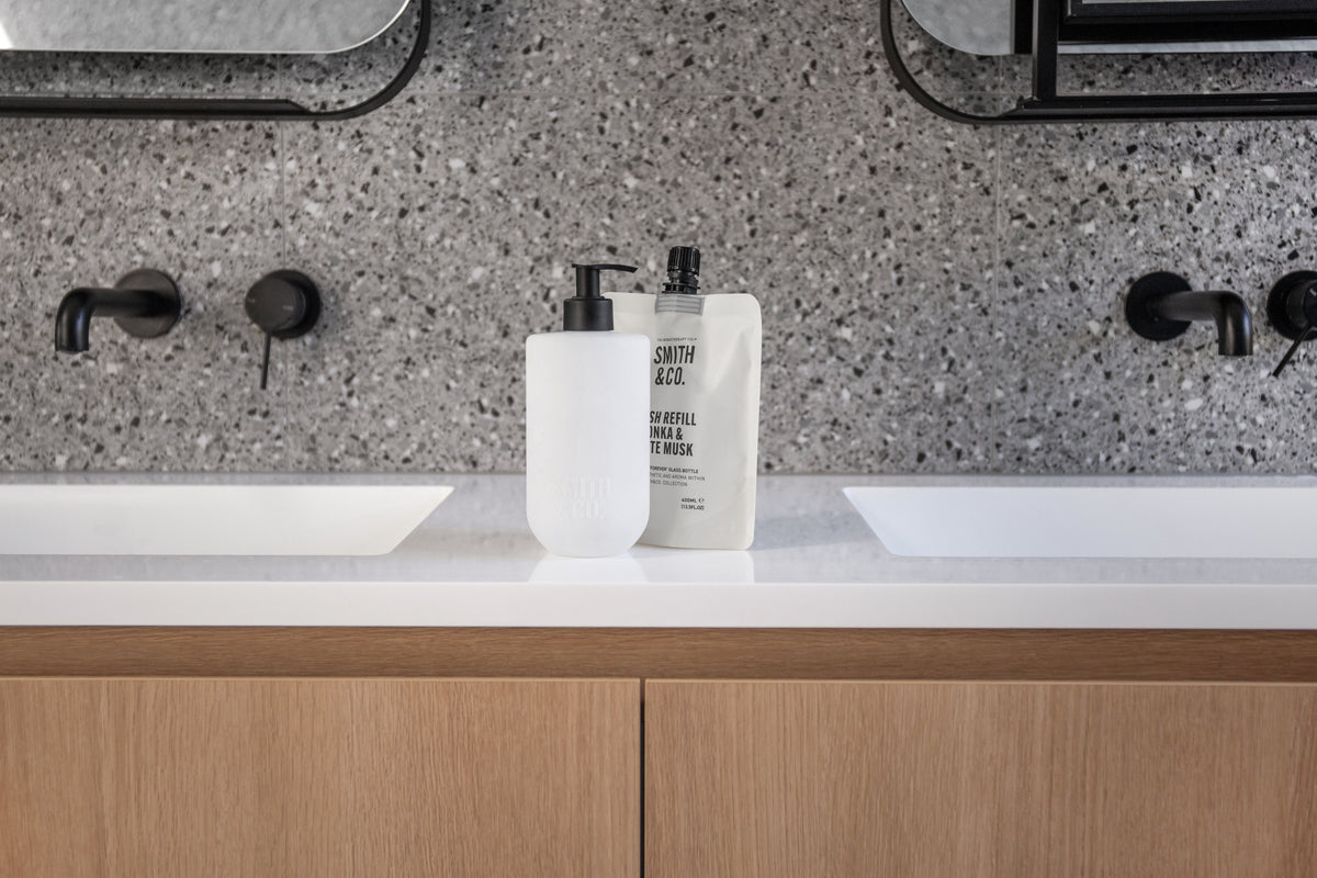 Frosted bottle and refill pouch between two basins on an oak double vanity, landscape view
