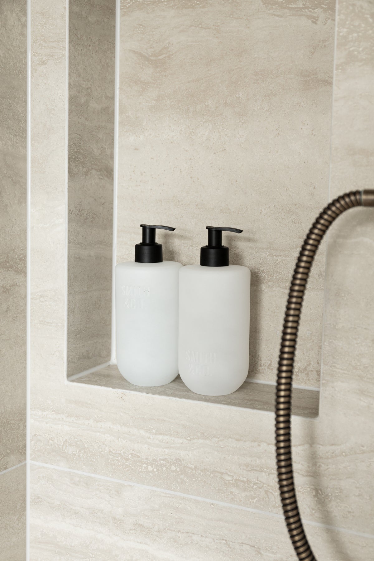Close view of two frosted pump bottles in a tiled shower niche with a brass hose curving past