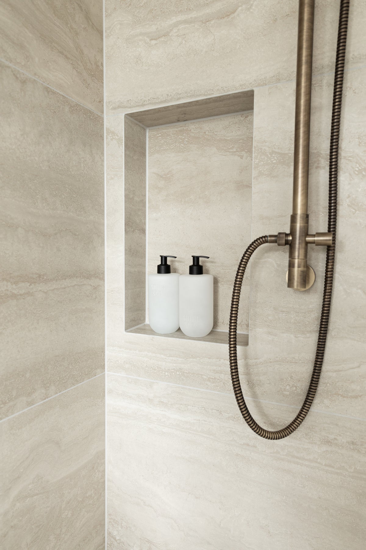 Two frosted pump bottles in a travertine-look shower niche beside a brushed brass shower hose