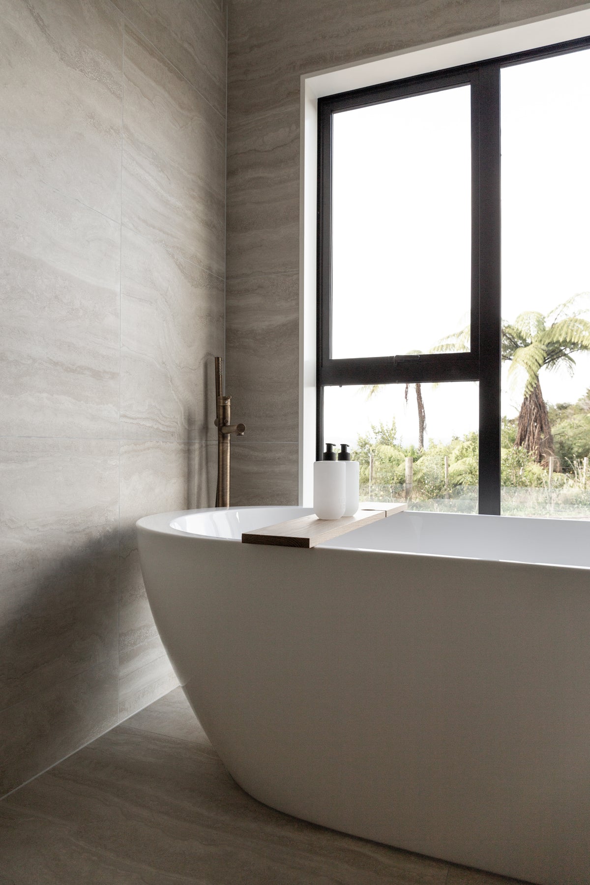 Freestanding bath by a tall window with palms outside and two small bottles on the rim